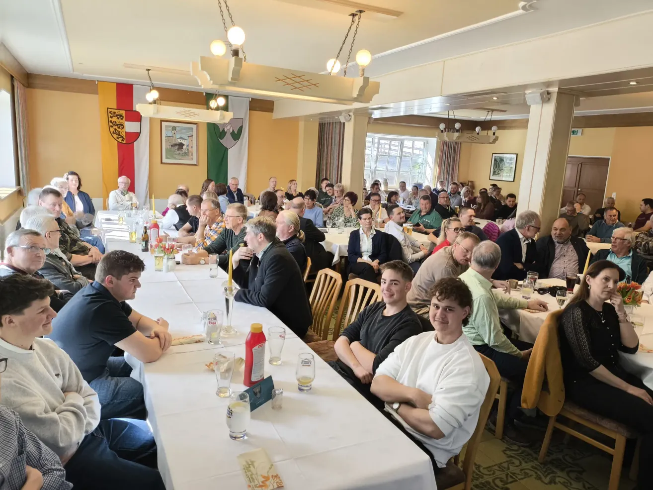 Eine große Gruppe von Menschen ist in einem Bankettsaal versammelt, an langen Tischen sitzend. Der Raum ist mit Flaggen und Bildern an den Wänden dekoriert. Einige unterhalten sich, während andere umherschauen. Verschiedene Gegenstände wie Flaschen, Gläser und Blumen stehen auf den Tischen.