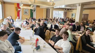 Eine große Gruppe von Menschen ist in einem Bankettsaal versammelt, an langen Tischen sitzend. Der Raum ist mit Flaggen und Bildern an den Wänden dekoriert. Einige unterhalten sich, während andere umherschauen. Verschiedene Gegenstände wie Flaschen, Gläser und Blumen stehen auf den Tischen.