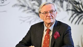 Ein älterer Mann in Anzug und Brille sitzt nachdenklich vor einem weißen Hintergrund mit Baumdesign.