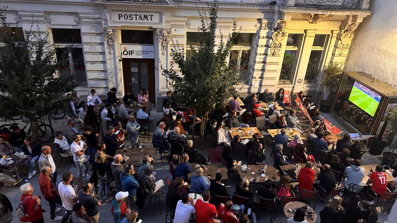 An outdoor dining area with many people sitting around tables. The building in the background has a sign reading 'Postamt' and 'OIF'.