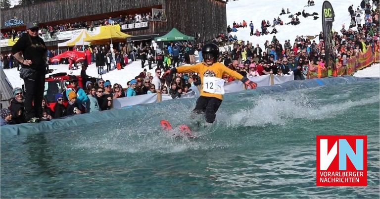 Ein Skifahrer springt ins Wasser mit der Nummer 12 auf dem Shirt, umgeben von Zuschauern an verschneiten Hängen mit Zelten und einem Gebäude im Hintergrund.
