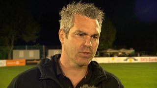 Ein Mann steht in einem Außenbereich, wahrscheinlich einem Fußballstadion. Er trägt eine schwarze Jacke und ein schwarzes Poloshirt. Er schaut nach vorne und sein Mund ist leicht geöffnet. Der Bereich hinter ihm ist verschwommen.