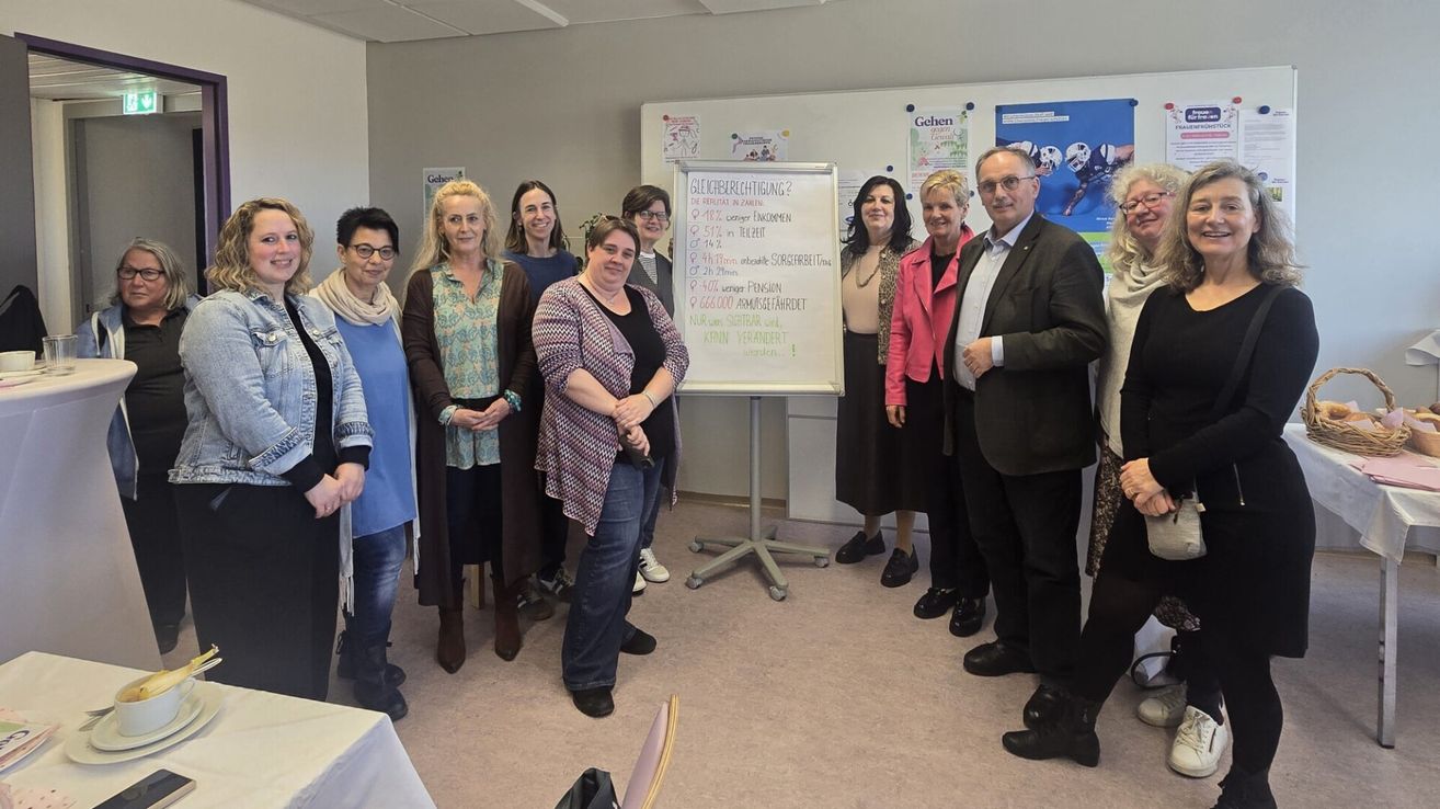 Several adults stand in a room, smiling, with a whiteboard behind them containing financial figures. Some wear glasses, jackets, and scarves. A man and woman stand closest to the whiteboard.