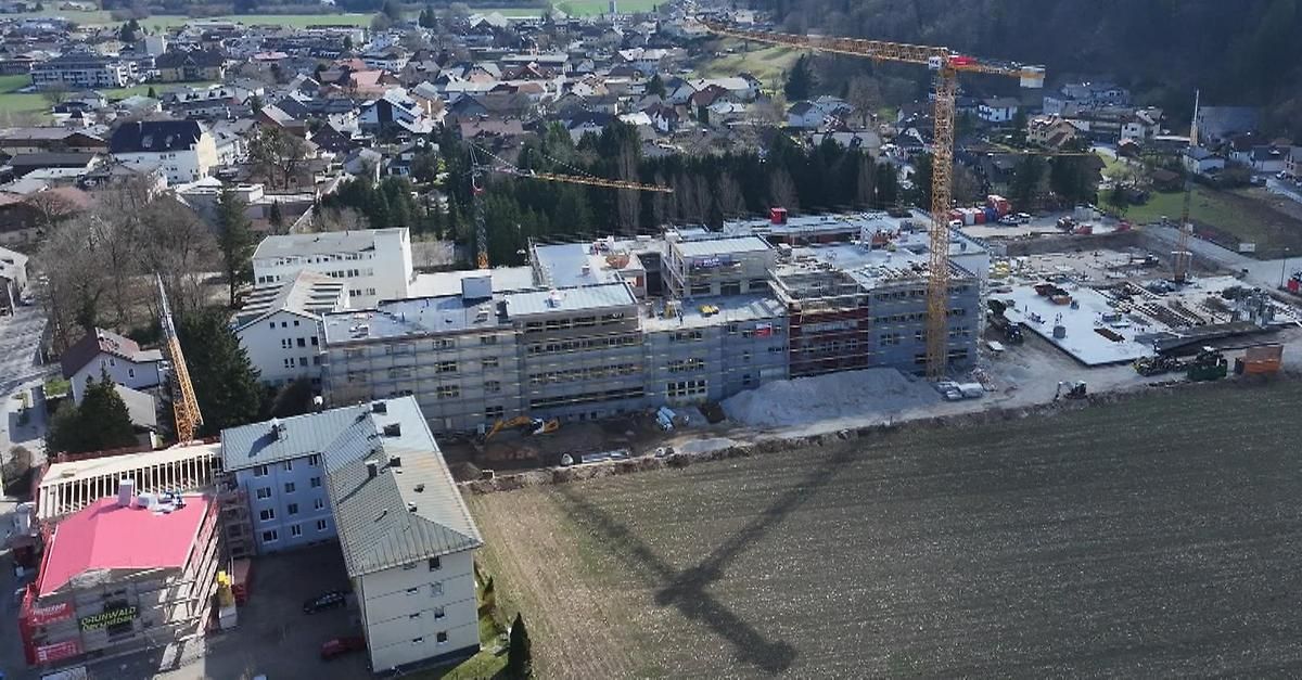 Luftaufnahme einer Baustelle mit im Bau befindlichen Gebäuden, Kränen und einem großen Feld davor.
