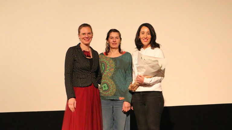 Drei Frauen stehen zusammen und lächeln für ein Foto. Eine hält einen Blumenstrauß, eine andere trägt eine Brille und eine gepunktete Jacke. Die dritte trägt ein grünes Oberteil und Jeans.