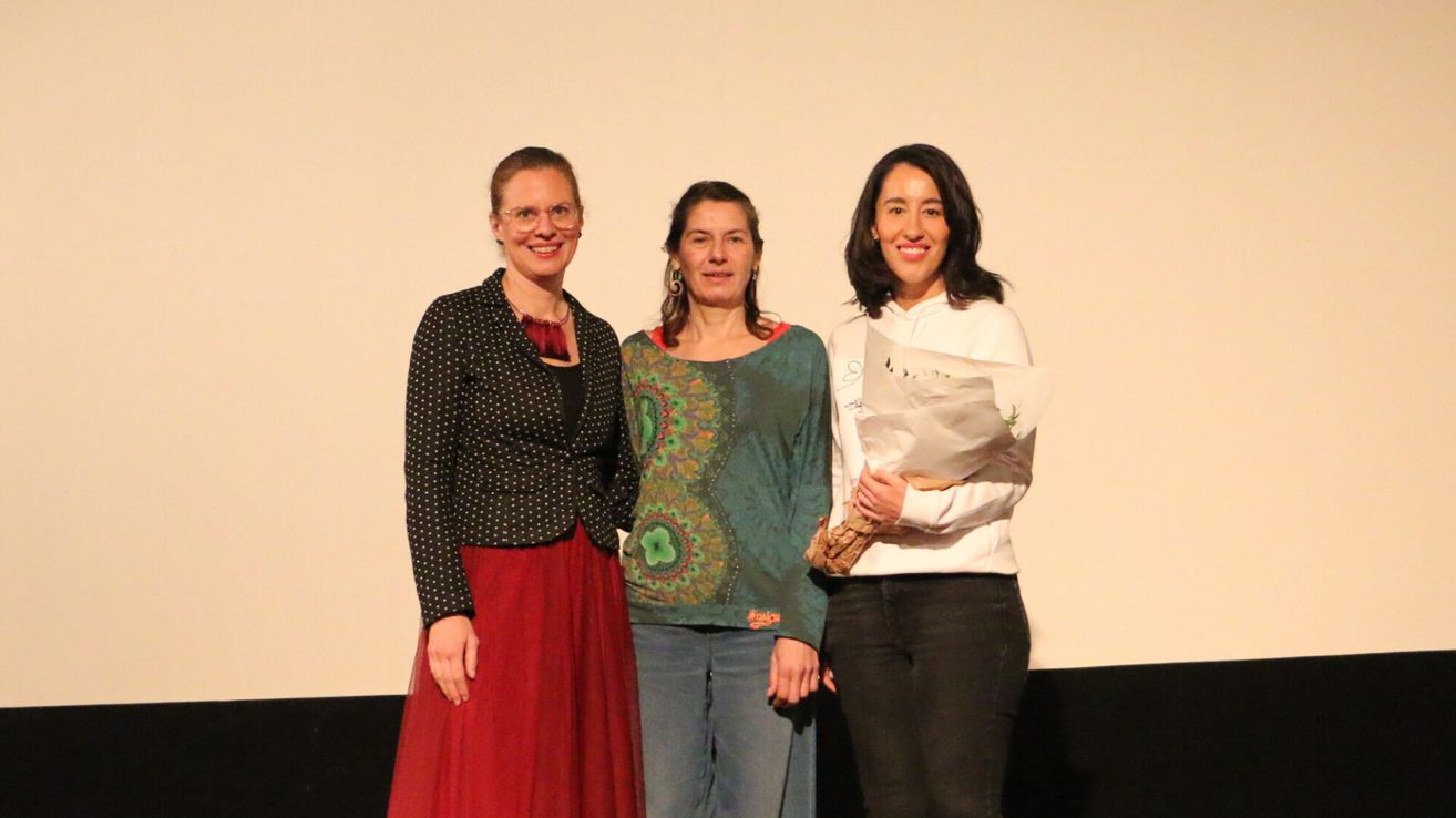 Drei Frauen stehen zusammen und lächeln für ein Foto. Eine hält einen Blumenstrauß, eine andere trägt eine Brille und eine gepunktete Jacke. Die dritte trägt ein grünes Oberteil und Jeans.