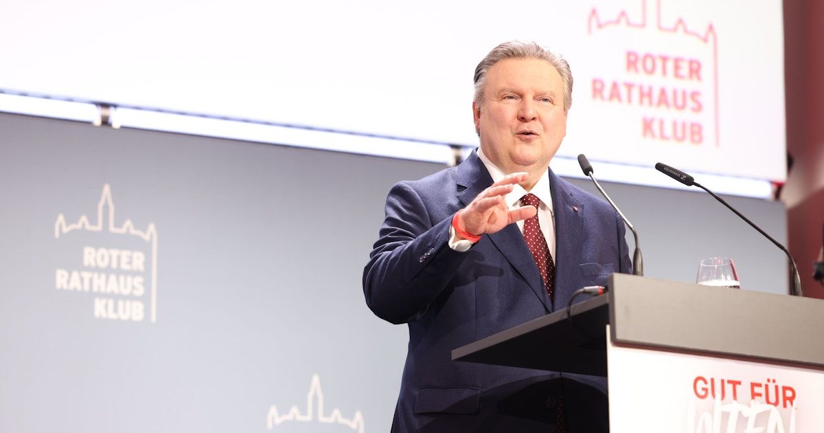 Ein Mann steht an einem Podium und spricht in ein Mikrofon. Er trägt einen dunkelblauen Anzug mit einer roten Krawatte. Hinter ihm zeigt eine Leinwand die Worte 'ROT RATHAU KL'.