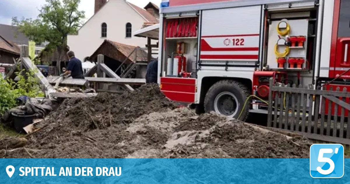 Ein Feuerwehrauto steht vor einem beschädigten Gebäude. Zwei Personen stehen in der Nähe des Bauwerks. Das Fahrzeug ist mit '122' gekennzeichnet. Unten befindet sich ein blauer Banner mit den Worten 'IN DER DRAU'.