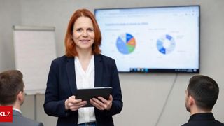 Eine Frau mit roten Haaren und einem Anzug lächelt, während sie ein Tablet hält und vor einem Bildschirm steht, auf dem ein Kreisdiagramm zu sehen ist.