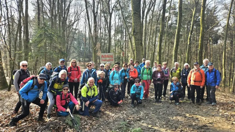 Eine Gruppe von Wanderern posiert für ein Foto im Wald. Sie tragen Winterkleidung und haben Rucksäcke dabei. Ein Schild im Hintergrund lautet 'Achtung Staatsgrenze'.