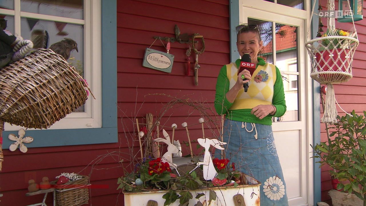 A woman stands in front of a red house, holding a microphone, and smiling. She wears a green and yellow sweater with a flower brooch. Nearby, a display of Easter decorations with flowers and bunny figures sits on a table.
