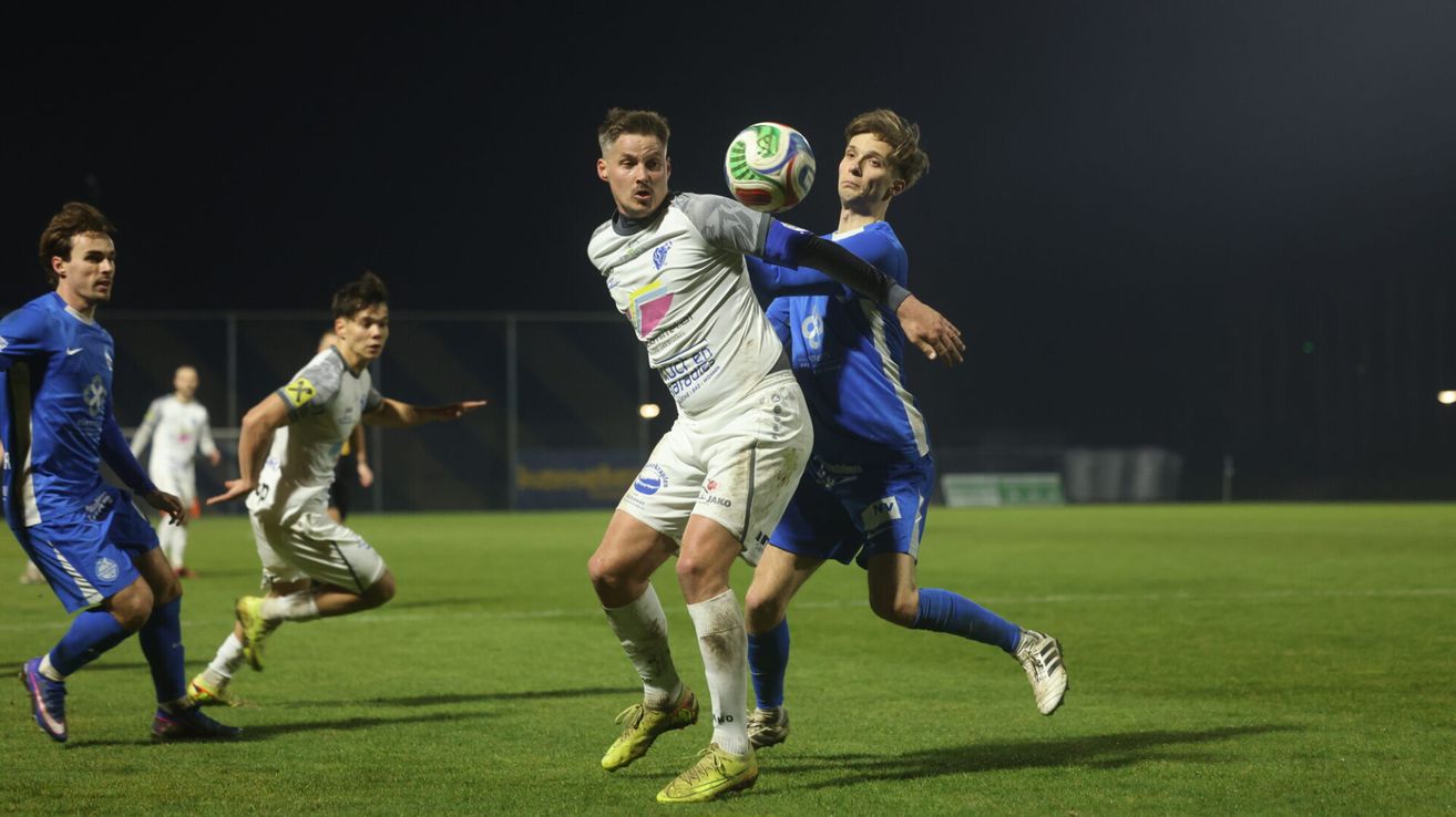 A soccer player in white is in mid-air, attempting to head a ball while another player in blue defends. The field is green with a fence in the background.