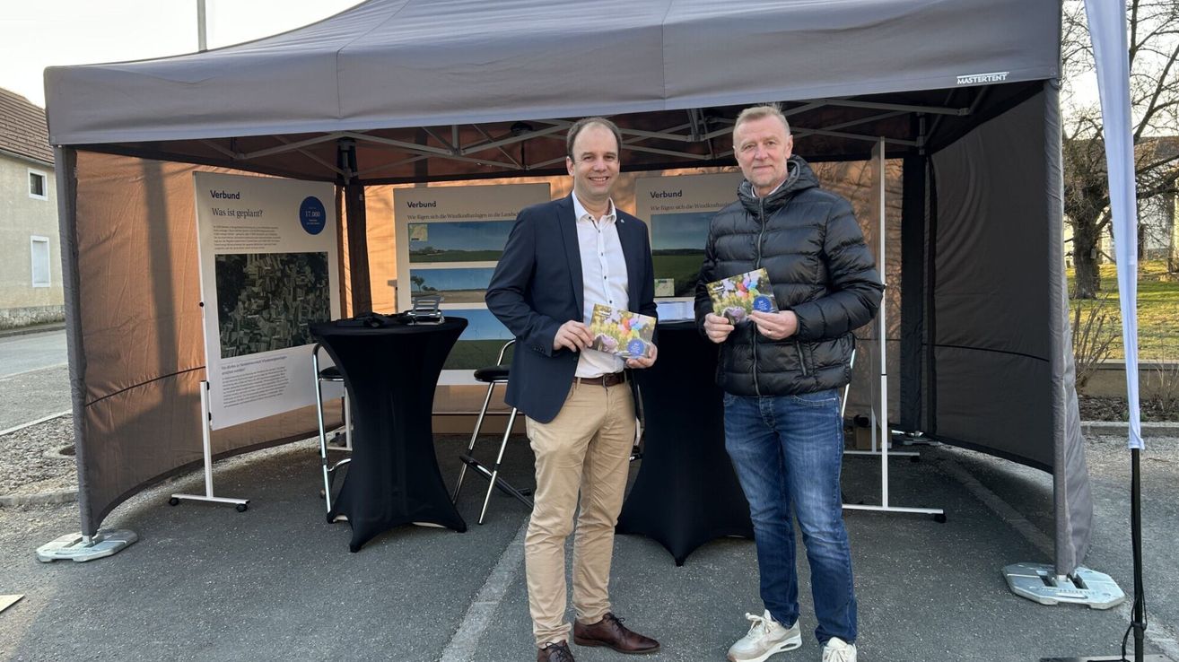 Zwei Männer stehen unter einem Zelt, lächeln und halten Bücher in den Händen. Hinter ihnen steht ein Tisch mit schwarzen Tischdecken und Hockern. Ein Poster mit dem Wort 'Verband' ist am Zelt befestigt. Der Boden ist grau.