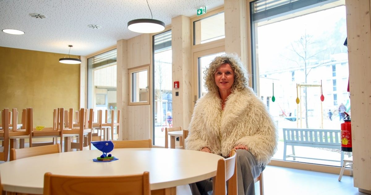 Eine Frau in einem weißen Pelzmantel sitzt auf einem Stuhl in einem Esszimmer mit einem runden Tisch. Auf dem Tisch steht ein blaues Spielzeug und ein Fenster zeigt einen Baum.