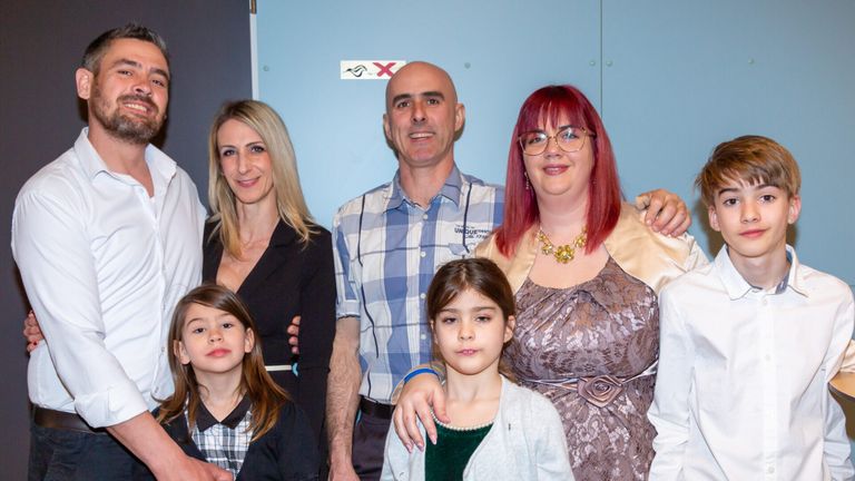 Ein Familienfoto mit vier Personen vor einer blauen Wand. Zwei Erwachsene und zwei Kinder. Die Erwachsenen lächeln.