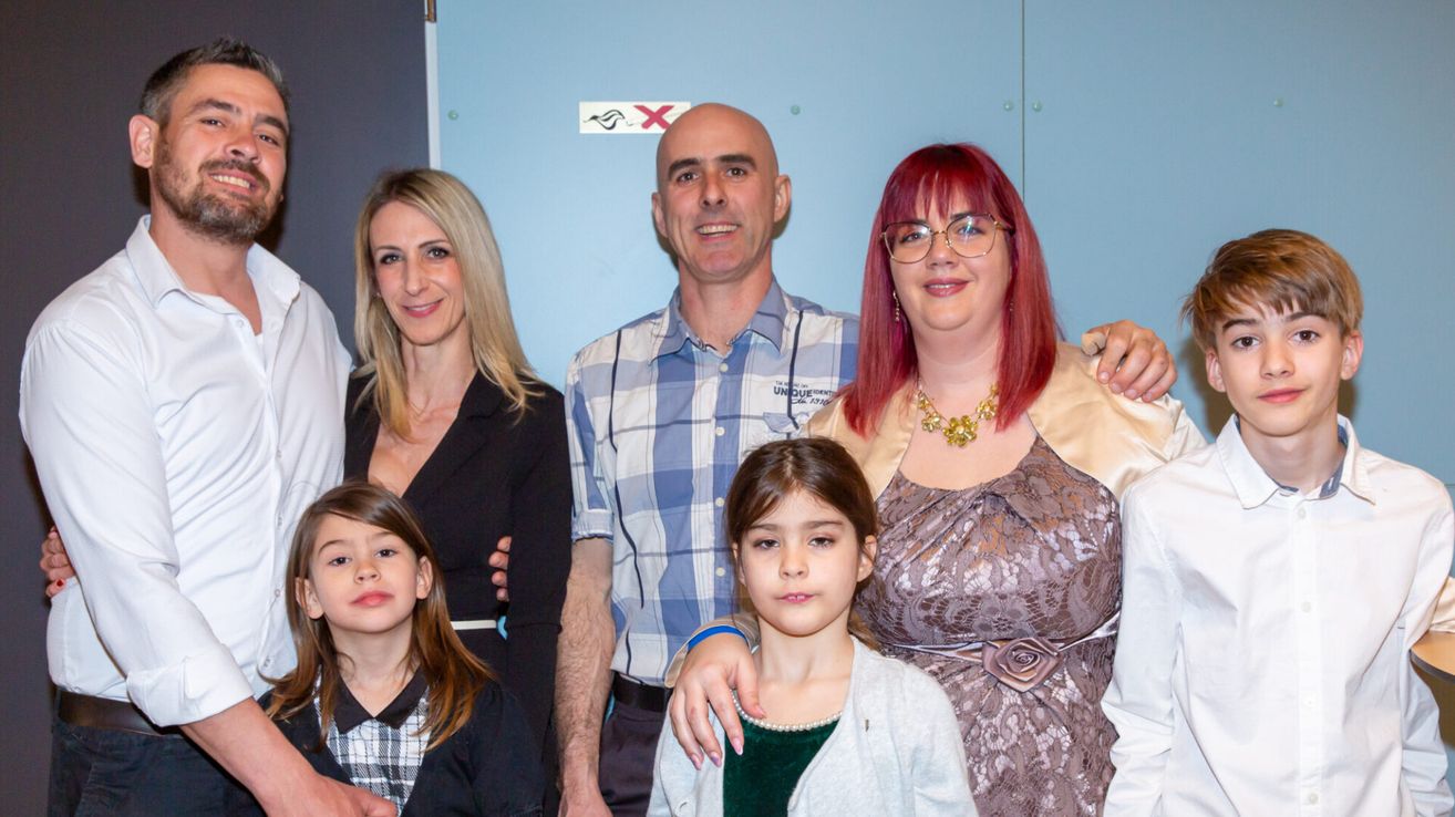 Ein Familienfoto mit vier Personen vor einer blauen Wand. Zwei Erwachsene und zwei Kinder. Die Erwachsenen lächeln.