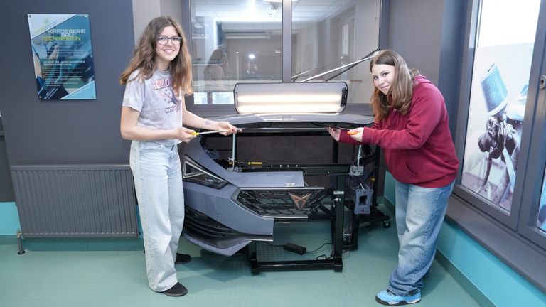 Zwei junge Frauen arbeiten in einer Werkstatt an einem Auto, der vordere Teil des Autos ist angehoben. Beide halten Werkzeuge und eine lächelt in die Kamera.