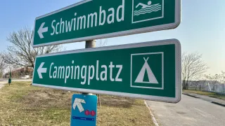 Two green road signs indicate 'Swimming Bad' and 'Campingplatz' with respective icons. The signs are mounted on a pole beside a grassy area. A small blue sign with hiking symbols is below.