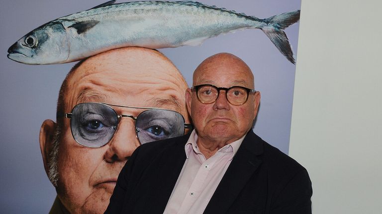 Ein älterer Mann mit Brille und Anzug steht vor einem Poster mit dem Gesicht eines Mannes, auf dessen Kopf ein Fisch sitzt.