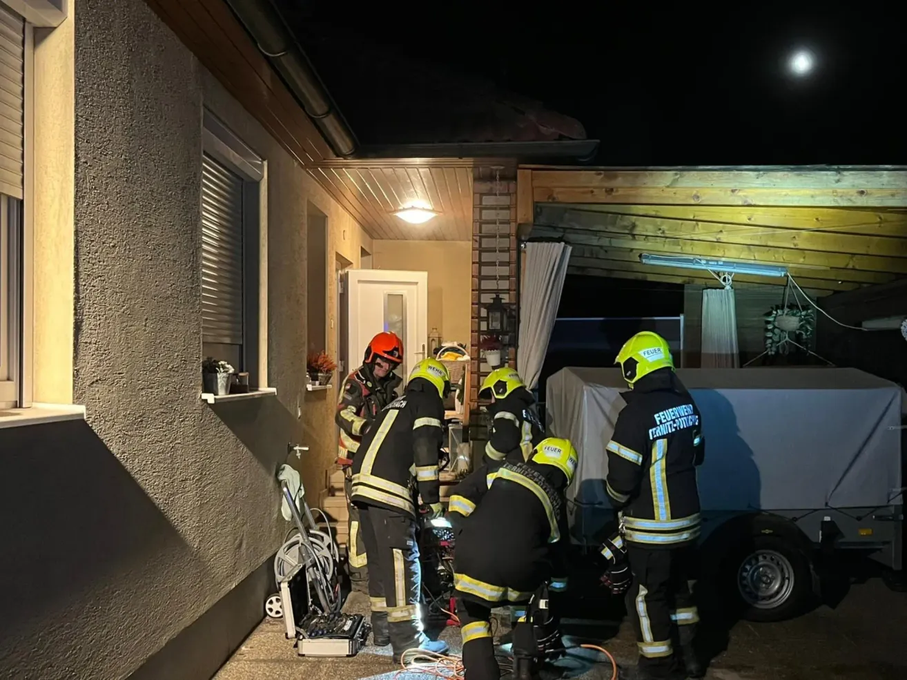 Firefighters in protective gear are working on a vehicle outside a house at night. One of them is bent over, possibly checking the vehicle. A car wheel is on the ground nearby.