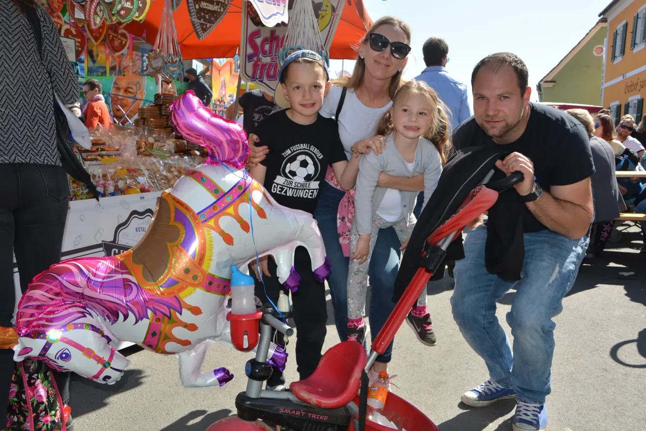 Eine Familie posiert für ein Foto vor einem farbenfrohen Pferdeballon und einer Karussellfahrt, wobei ein Mann einen Kinderwagen hält und ein anderer Mann im Hintergrund geht.