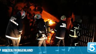 Firefighters in protective gear extinguish a building fire at night. The fire is intense, and the firefighters use hoses to combat the flames.