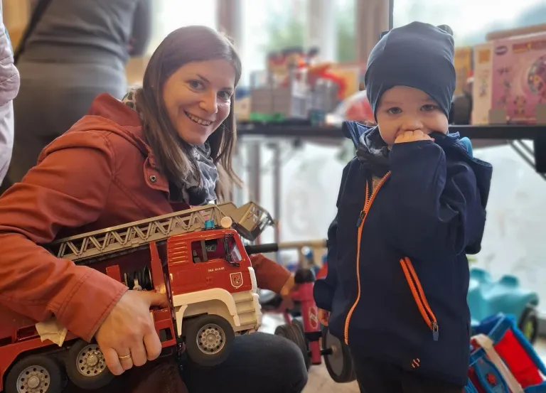 A woman is holding a toy fire truck and smiling. A young child in a blue coat with an orange zipper stands next to her. Behind them, various toys and a blurry background are visible.