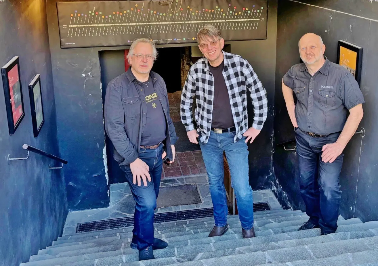 Three men stand on steps; one wears a checkered shirt, another a denim jacket with text, and the third a gray shirt. They smile, standing by a wall with a map.
