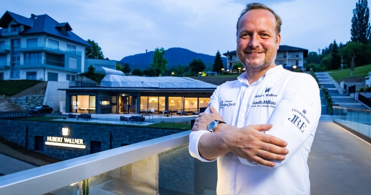 Ein lächelnder Mann steht mit verschränkten Armen auf einem Balkon und trägt ein weißes Kochoutfit. Hinter ihm steht ein großes Gebäude mit Glasdach und einer malerischen Aussicht auf Berge.