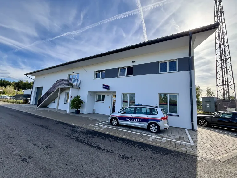 Ein Polizeiauto ist vor einem weißen Gebäude mit grauen Akzenten geparkt. Das Gebäude hat eine Treppe, die zu einem Balkon führt. Eine Topfpflanze befindet sich an der Seite.