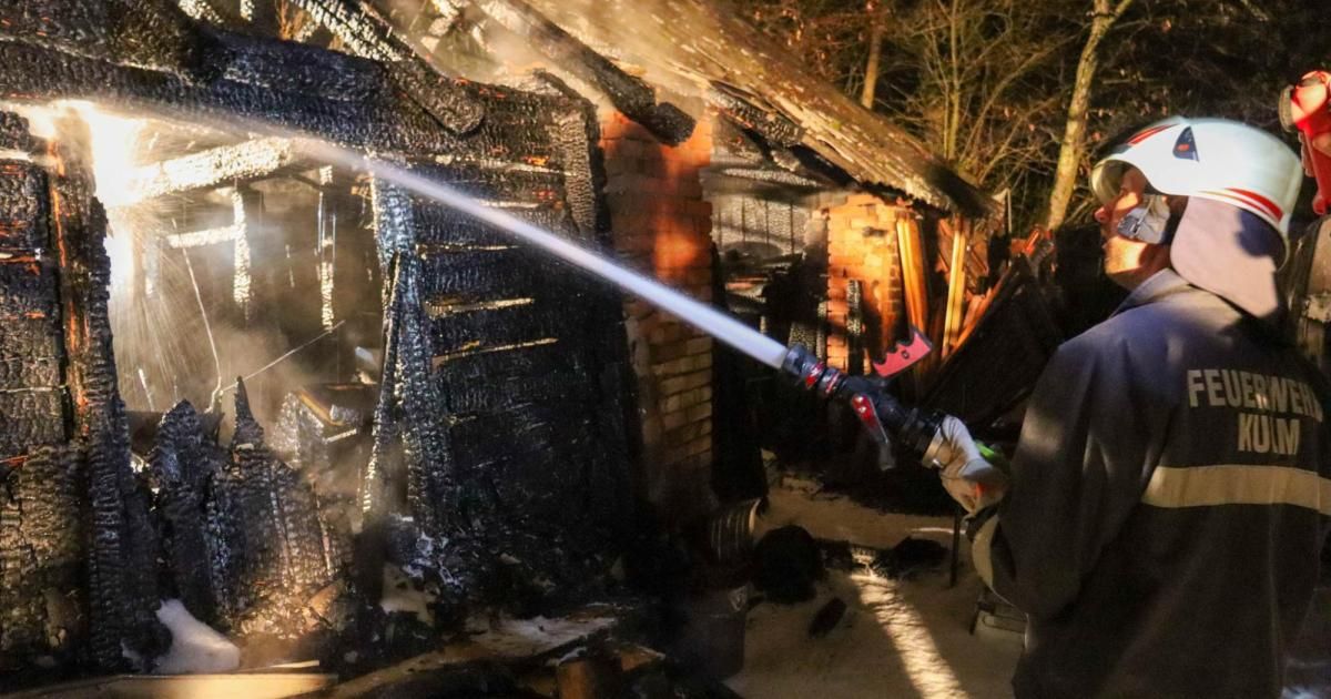 A firefighter sprays water on a burnt-out building with a damaged roof and charred walls, surrounded by debris and trees.