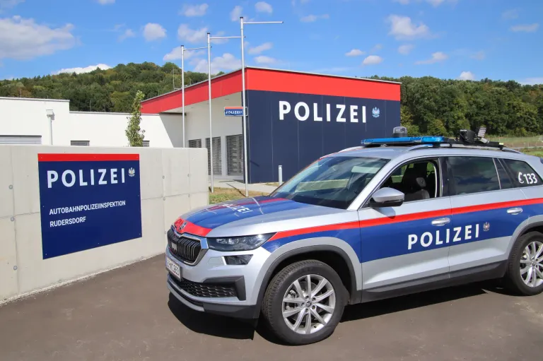Ein Polizei-SUV ist vor einem Gebäude mit dem Wort 'POLIZEI' darauf geparkt. Der SUV hat das Wort 'POLIZEI' an seiner Seite und auf dem Dach. Es gibt ein Schild mit dem Text 'POLIZEIINSPEKTION' auf der linken Seite. Bäume und ein Berg sind im Hintergrund.