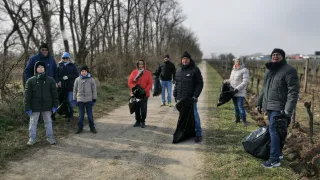 A group of people wearing winter clothing are standing on a dirt path, holding garbage bags. They are in a rural area with trees and grass.