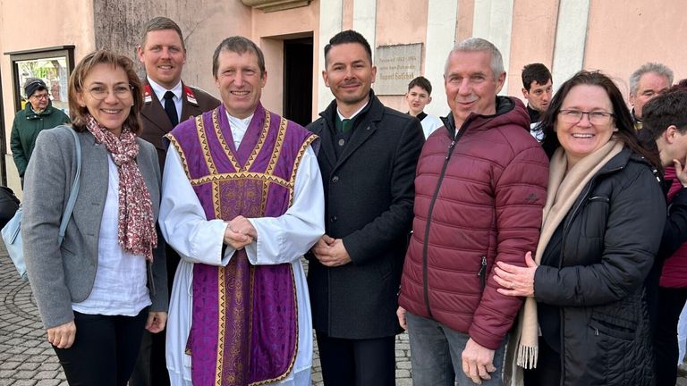 Eine Gruppe von Männern steht vor einem Gebäude mit einem Schild, einer in geistlicher Kleidung, zwei in Wintermänteln und ein anderer in Uniform, alle lächeln.