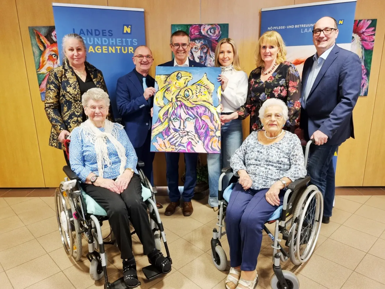 Eine Gruppe von Menschen steht und sitzt in einem Raum. Zwei ältere Frauen sind in Rollstühlen und lächeln. Ein Mann hält ein farbenfrohes Gemälde eines Gesichts. Der Raum hat Banner an der Wand und einen gefliesten Boden.