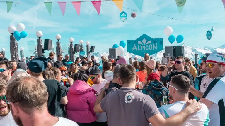 Eine große Menschenmenge versammelt sich beim Alpicon-Festival mit bunten Flaggen, Lautsprechern und Luftballons. Die Leute machen Fotos, genießen Getränke und unterhalten sich vor einer Bergkulisse.