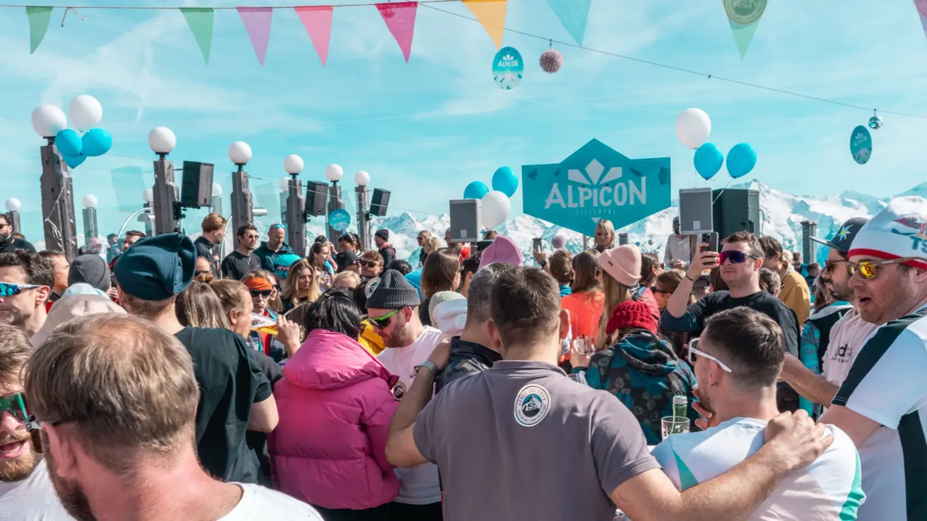 Eine große Menschenmenge versammelt sich beim Alpicon-Festival mit bunten Flaggen, Lautsprechern und Luftballons. Die Leute machen Fotos, genießen Getränke und unterhalten sich vor einer Bergkulisse.
