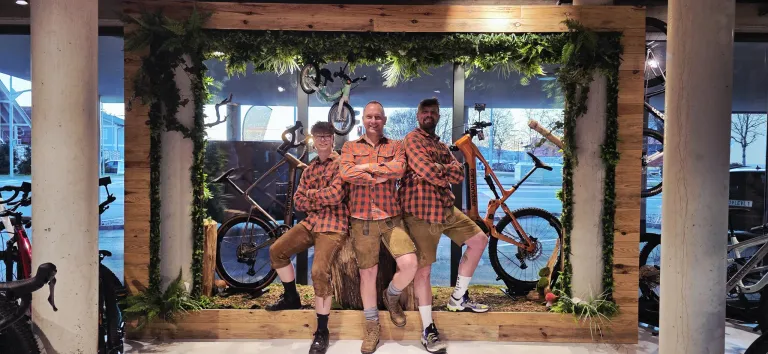 Three men in matching plaid shirts and shorts pose for a photo in front of bicycles, with one man crossing his arms and another sitting on a tree stump.