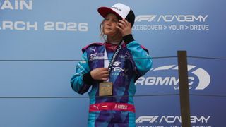 A young female racing driver stands on the podium, wearing a blue racing suit with a gold medal around her neck. The backdrop features a blue wall with white text and logos.