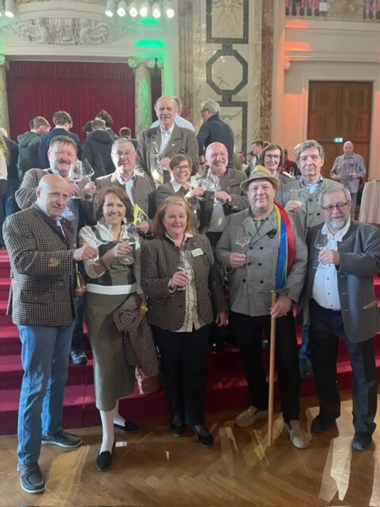 Eine Gruppe von Erwachsenen, sowohl Männer als auch Frauen, lächeln und halten Weingläser in der Hand, wahrscheinlich feiern sie ein Ereignis in einem Saal mit roten Teppichtreppen und einem roten Vorhang.