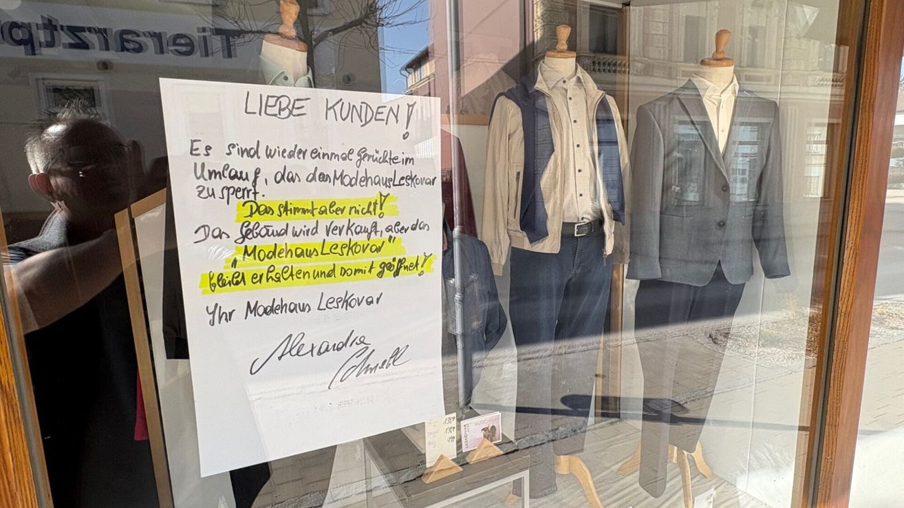 A handwritten sign is placed in front of a clothing store window. It reads 'LIEBE KUNDEN' in large letters, followed by a message in German about the stock. Mannequins with clothing are visible in the window.