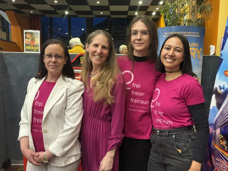 Vier Frauen stehen zusammen und lächeln. Sie tragen passende rosafarbene T-Shirts mit 'freier freiraum' darauf. Im Hintergrund ist ein blauer Banner und eine gemusterte Decke zu sehen.