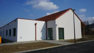 A modern white building with a red roof and red trim is under construction. It has two glass doors and a red pipe on the wall.