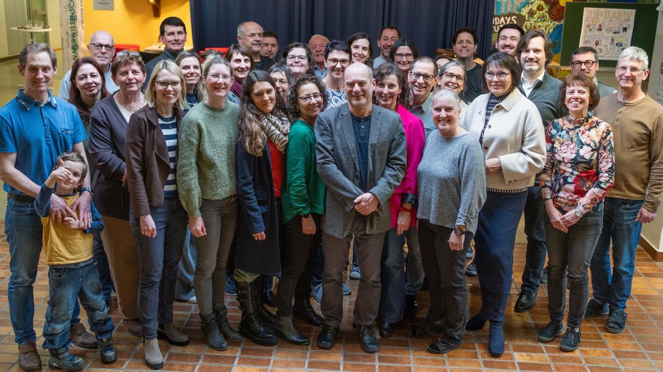 Eine Gruppe von Menschen posiert für ein Foto. Sie tragen Winterkleidung. Der Raum hat einen gefliesten Boden und eine schwarze Vorhang im Hintergrund.