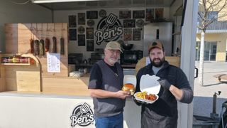 Two men pose with food at a Ben's BBQ stand, one holding a burger and the other holding a box of fries and chicken wings.