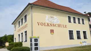 A yellow building with the words 'Volksschule' and a logo above a phone booth on the side. It has a red roof and white trim.
