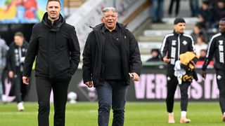 Drei Männer gehen auf einem Fußballfeld. Einer hält ein Handy und scheint zu telefonieren. Ein anderer trägt einen gelben Handschuh.