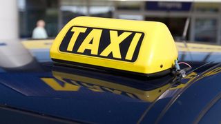 Ein Nahaufnahme von einem gelben Taxischild auf einem dunkelblauen Autodach, das sich auf der Oberfläche des Autos spiegelt.