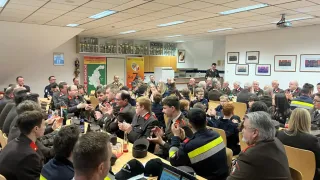 Ein Raum voller Menschen in Uniformen, die applaudieren. Laptops, Flaschen und Mützen liegen auf dem Tisch. Bilderrahmen hängen an der Wand. Ein Mann steht vor einer Tafel.