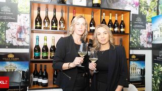 Two women stand before a wine display, each holding a glass of white wine. Behind them, shelves filled with various bottles of wine are visible. The women wear black outfits and appear to be smiling.